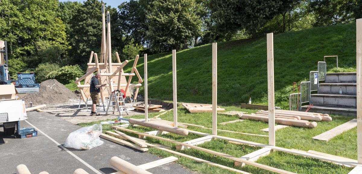 Baustelle mit Holzpfosten und -balken; Arbeiter montiert Teile einer Kletteranlage auf einer grünen Wiese.