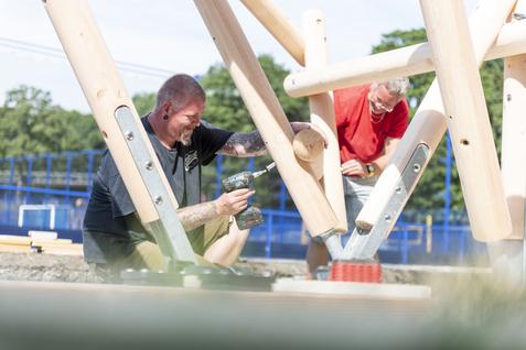 Zwei Männer montieren eine Holzstruktur im Freien. Einer benutzt einen Akkuschrauber, der andere hält ein Bauteil.