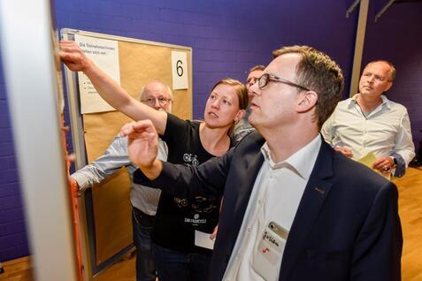Eine Gruppe von vier Personen steht vor einem Präsentationsboard und diskutiert, während eine Person auf das Board zeigt.
