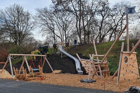 Spielplatz mit Rutsche, Klettergerüst und Schaukeln, umgeben von Bäumen und Bauarbeiten im Hintergrund.