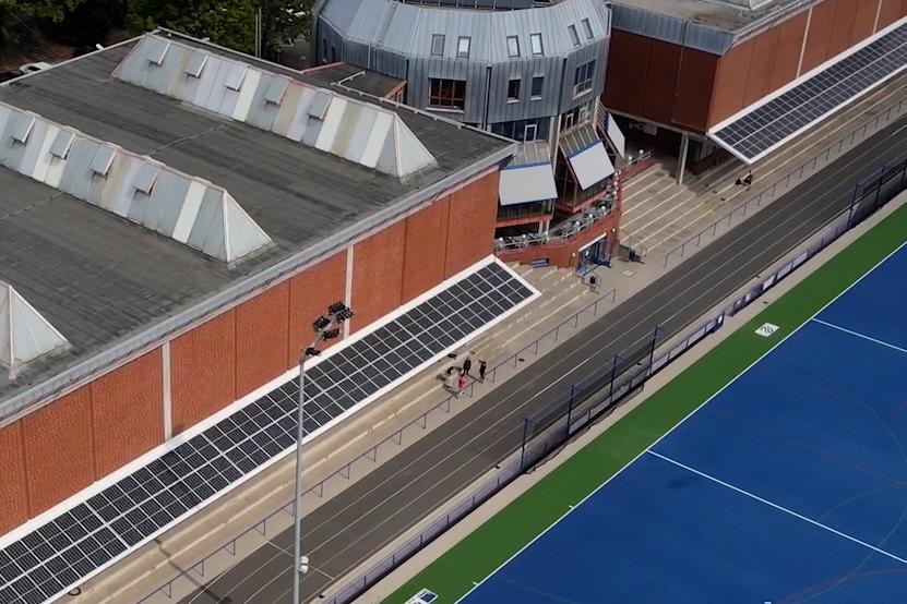 Luftaufnahme eines Sportkomplexes mit einem Hockeyfeld und modernen Sporthallen im Hintergrund.