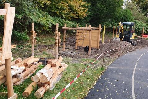 Holzspielgeräte und Baumstämme liegen an einem unvollendeten Spielplatz neben einem Radweg im Freien.