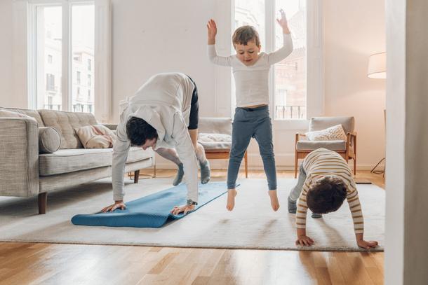 Familienmitglied springt freudig auf einem Teppich, w&auml;hrend zwei andere durch Sport&uuml;bungen auf dem Boden aktiv sind.