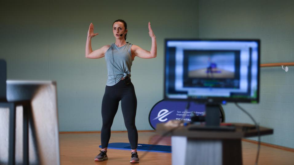 Fitnesstrainerin mit Headset führt Übungen vor Webcam in einem Trainingsraum durch. Computer zeigt Online-Teilnehmer.