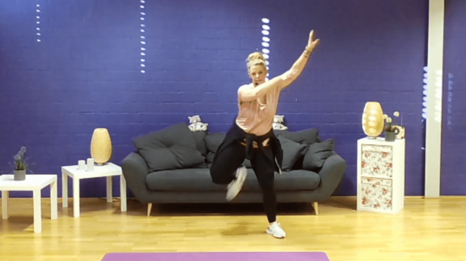 Frau in Sportkleidung führt eine dynamische Übung vor einem Sofa und lila Wand aus. Yogamatte liegt auf dem Boden.