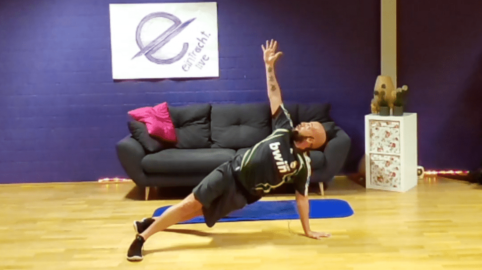 Mann in Sportkleidung führt eine seitliche Plank-Übung auf einer Matte in einem Wohnzimmer aus.