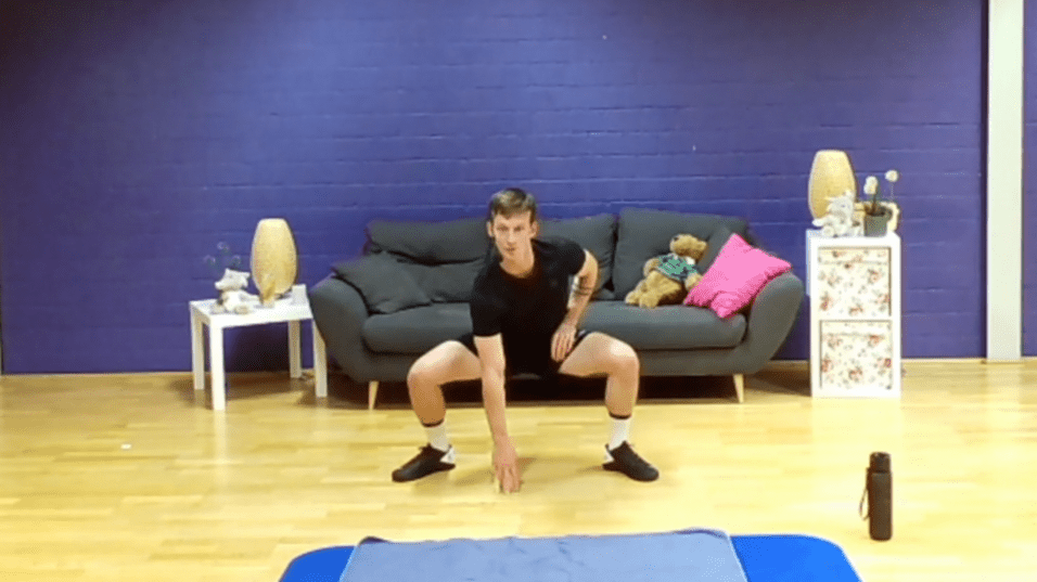 Junger Mann in Sportkleidung beugt sich vor einem blauen Gymnastikmatte im Wohnzimmer mit Sofa und Deko.