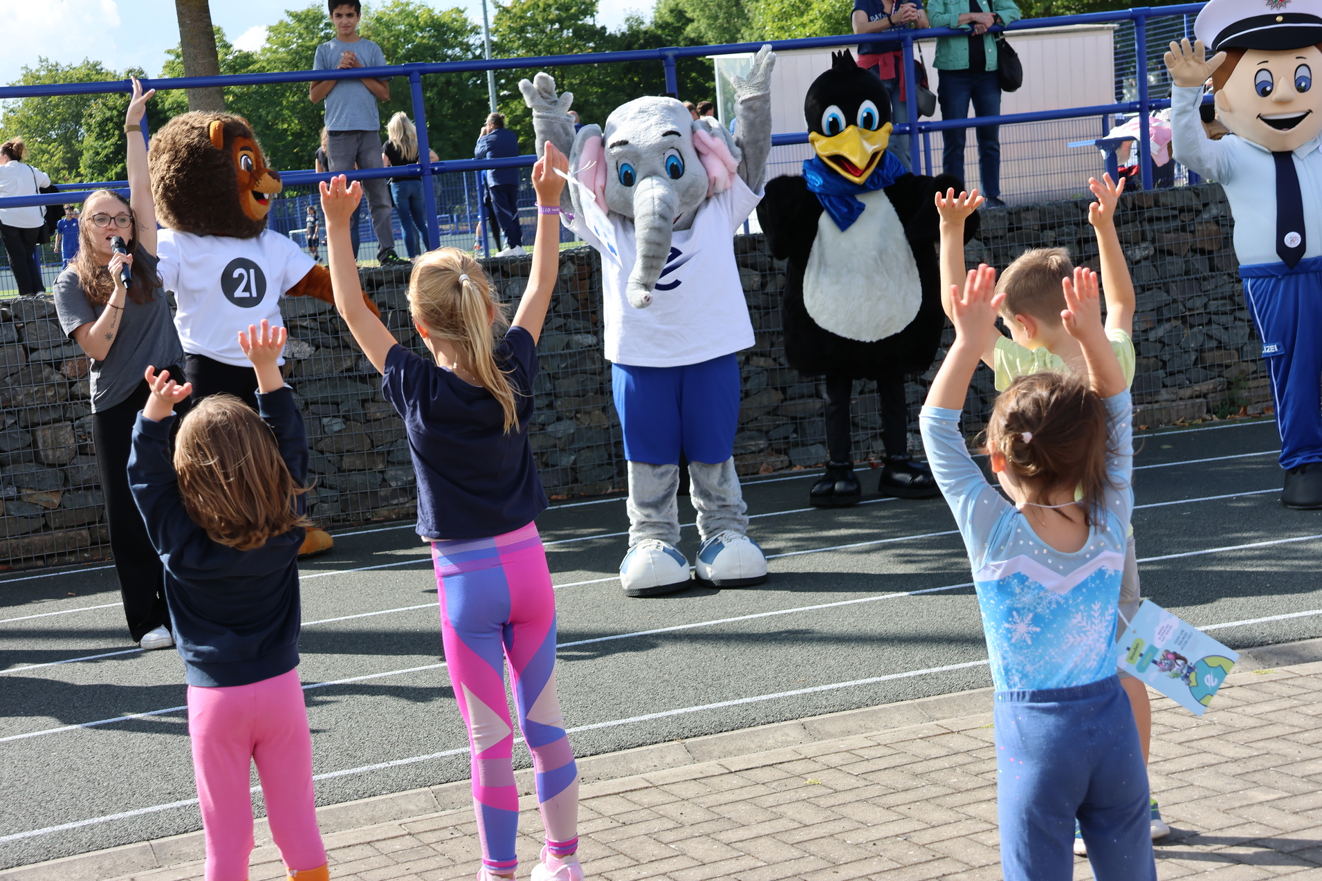Kinder stehen mit erhobenen H&auml;nden vor Maskottchen, das einen Elefanten darstellt, bei einer Veranstaltung im Freien.