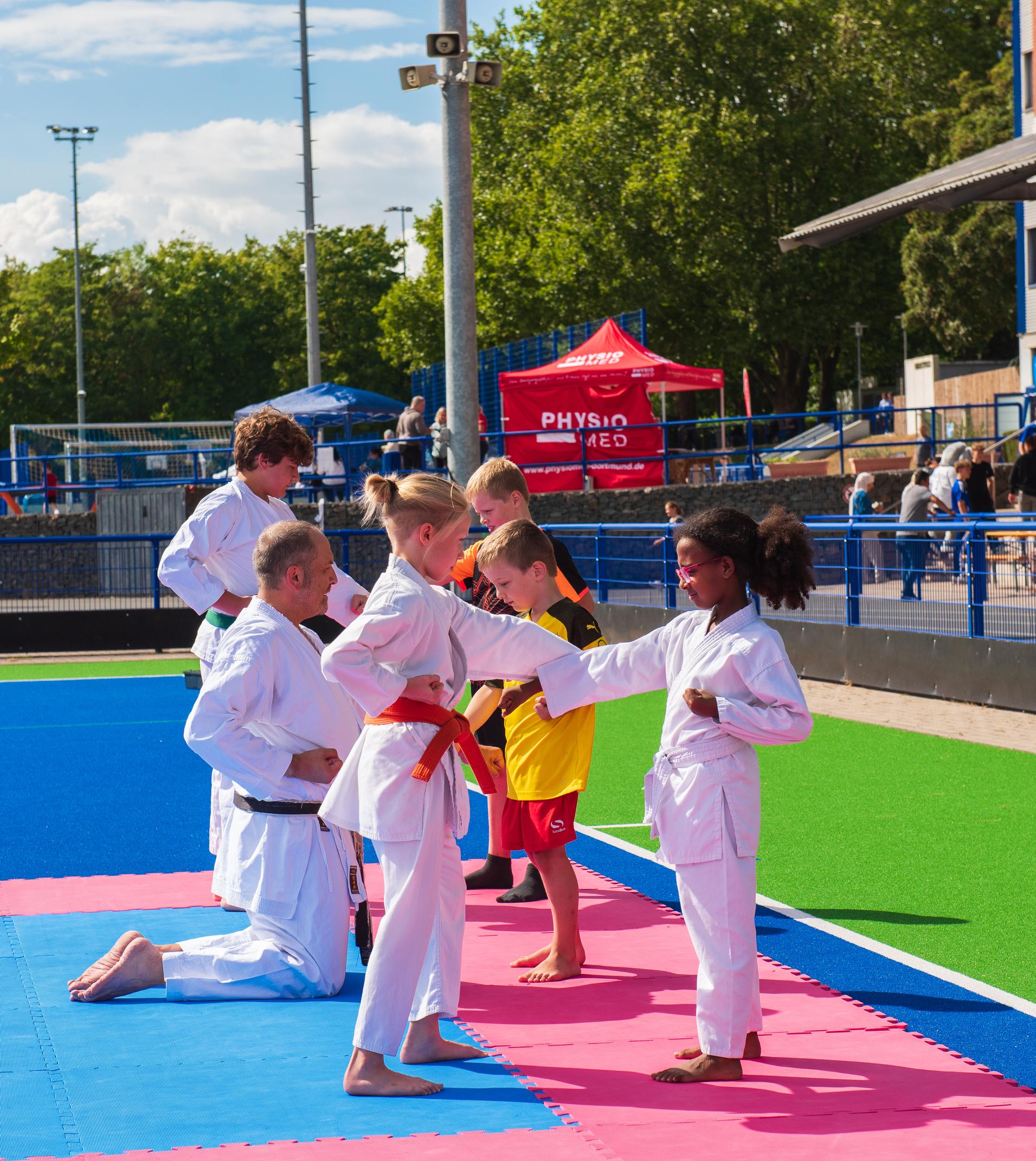 Kinder in Karate-Anz&uuml;gen &uuml;ben Techniken auf einer farbigen Matte, w&auml;hrend Trainer Anweisungen geben.