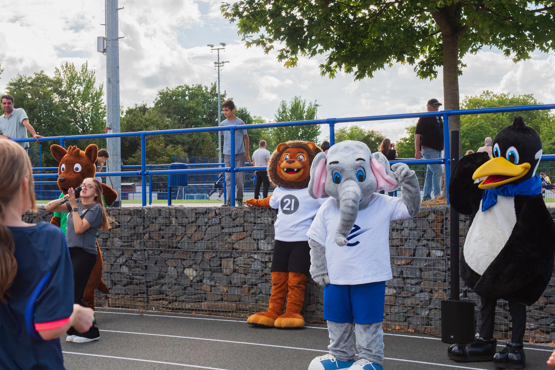 Vier bunte Tiermaskottchen stehen auf einem Sportplatz, umgeben von Menschen und B&auml;umen, und posieren f&uuml;r die Kamera.