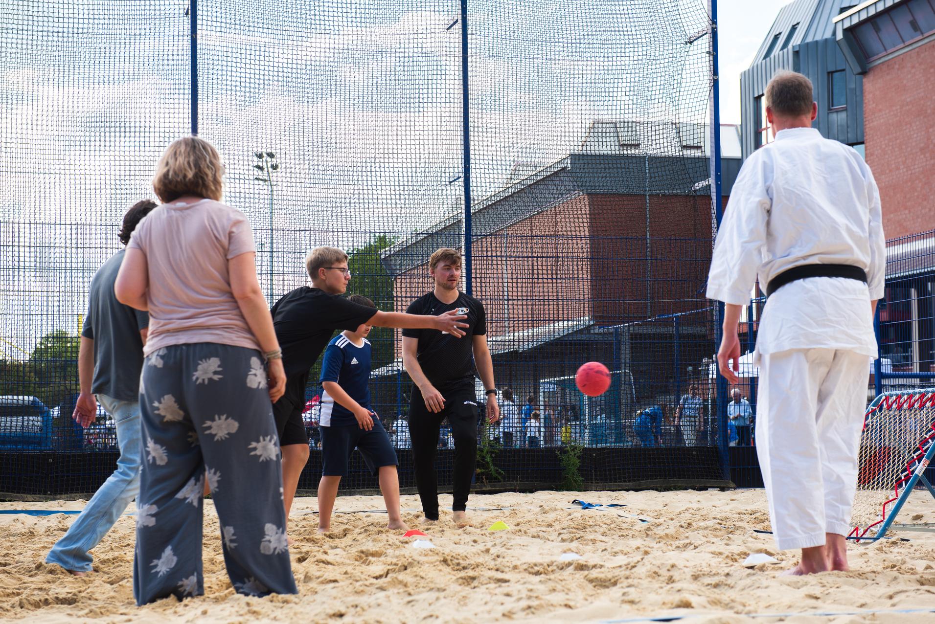 Gruppe von Menschen spielt im Sand eine Ballspiel-Variante auf einem Sportplatz, im Hintergrund ein Karateka.