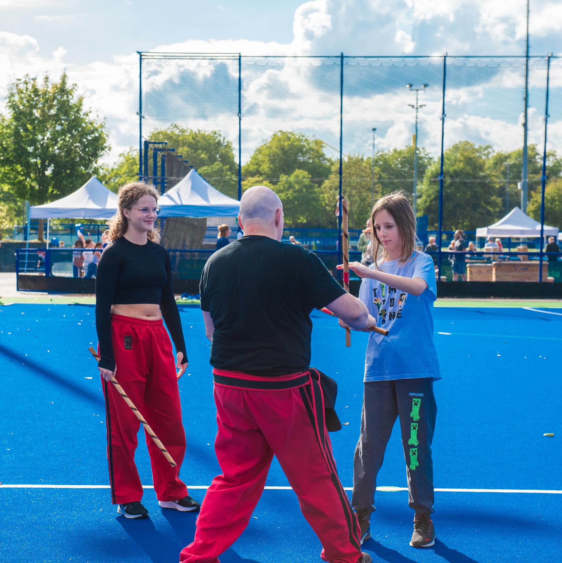 Drei Personen &uuml;ben mit St&ouml;cken auf einem blauen Sportplatz, im Hintergrund sind Zelte und B&auml;ume sichtbar.