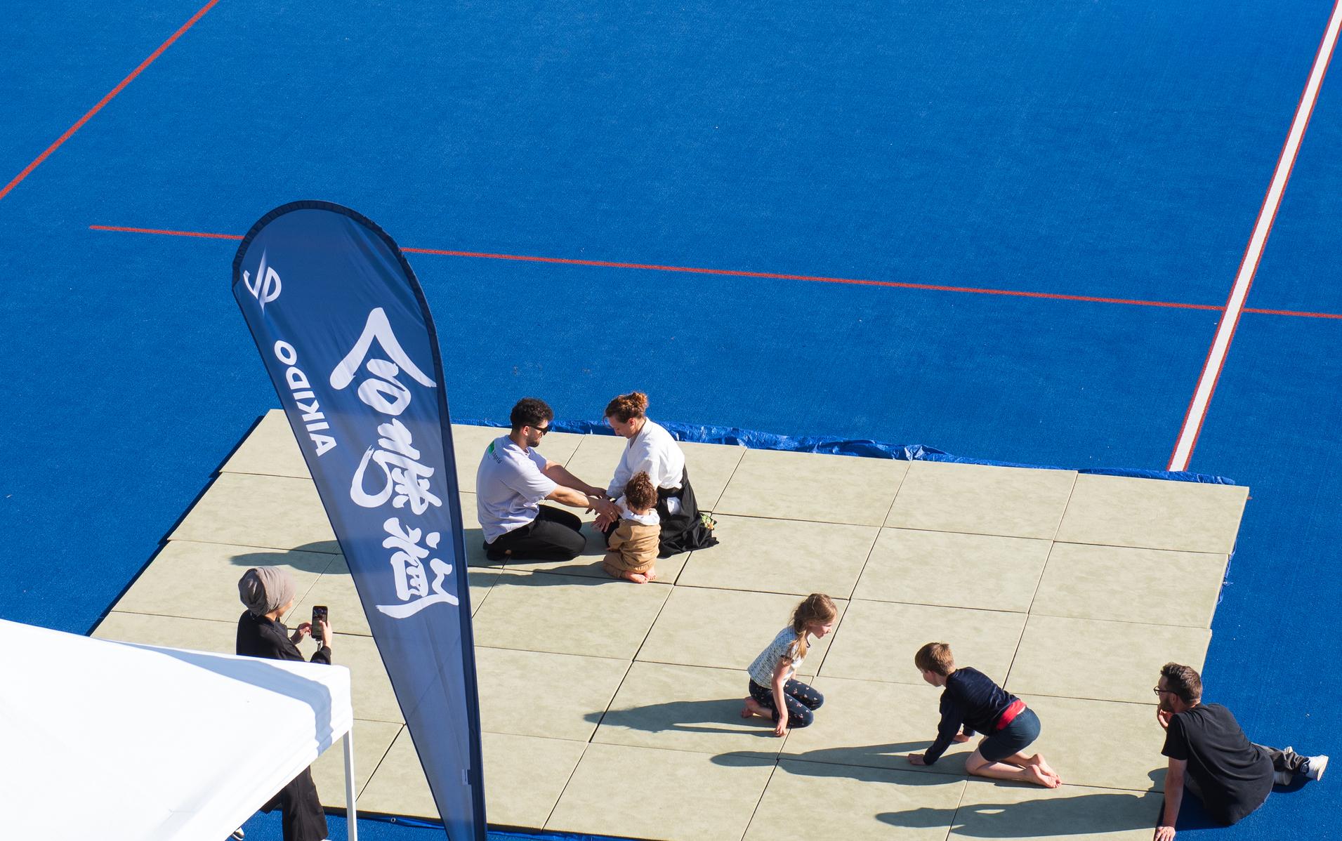 Gruppierung von Kindern und Erwachsenen auf einer Matte; im Hintergrund ein blaues Sportfeld und ein Fahne.
