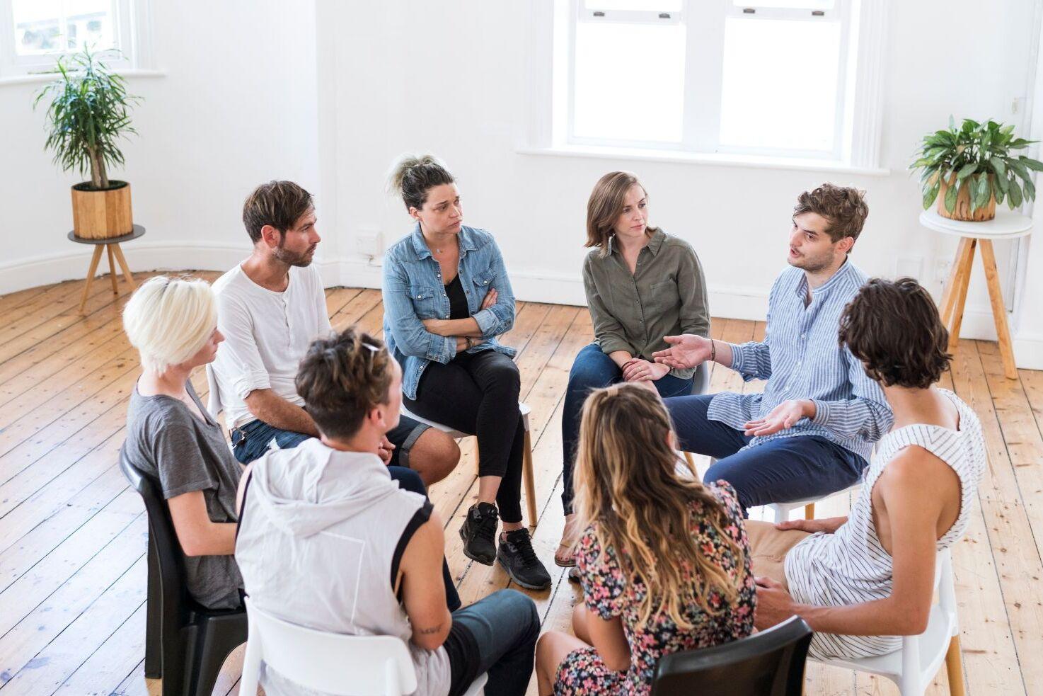 Gruppe von neun Personen in einem hellen Raum, die in einem Kreis miteinander diskutieren.