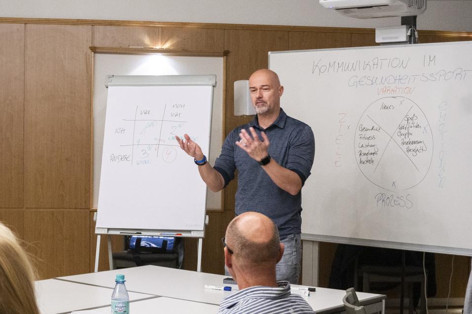 Mann spricht vor Publikum in einem Seminarraum, w&auml;hrend eine Flipchart und ein Projektor im Hintergrund sichtbar sind.