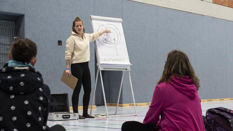Lehrerin erklärt eine Übung vor sitzenden Teilnehmern auf Yogamatten in einer Sporthalle.