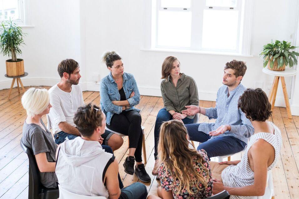 Gruppe von neun Personen in einem hellen Raum, die in einem Kreis miteinander diskutieren.