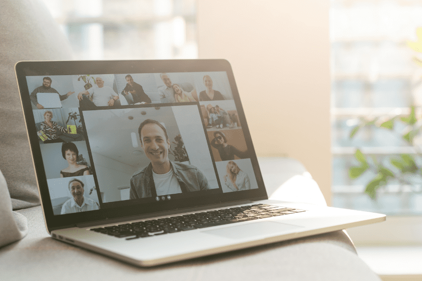 Laptop mit einer Videokonferenz, auf dem Gesichter von mehreren Teilnehmern und ein L&auml;chelnder im Vordergrund zu sehen sind.