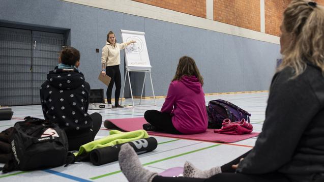 Lehrerin erklärt eine Übung vor sitzenden Teilnehmern auf Yogamatten in einer Sporthalle.