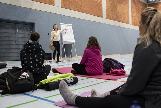 Lehrerin erkl&auml;rt eine &Uuml;bung vor sitzenden Teilnehmern auf Yogamatten in einer Sporthalle.
