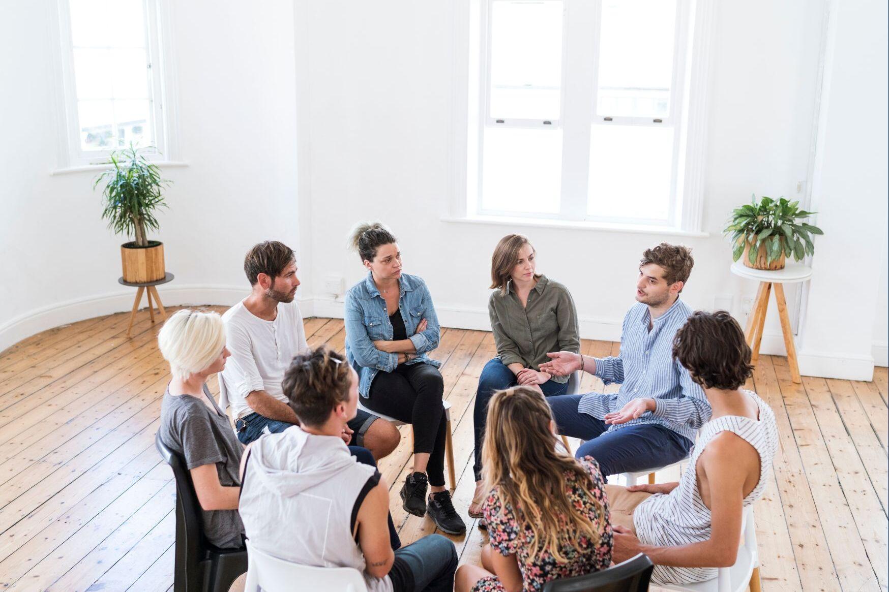 Gruppe von neun Personen in einem hellen Raum, die in einem Kreis miteinander diskutieren.