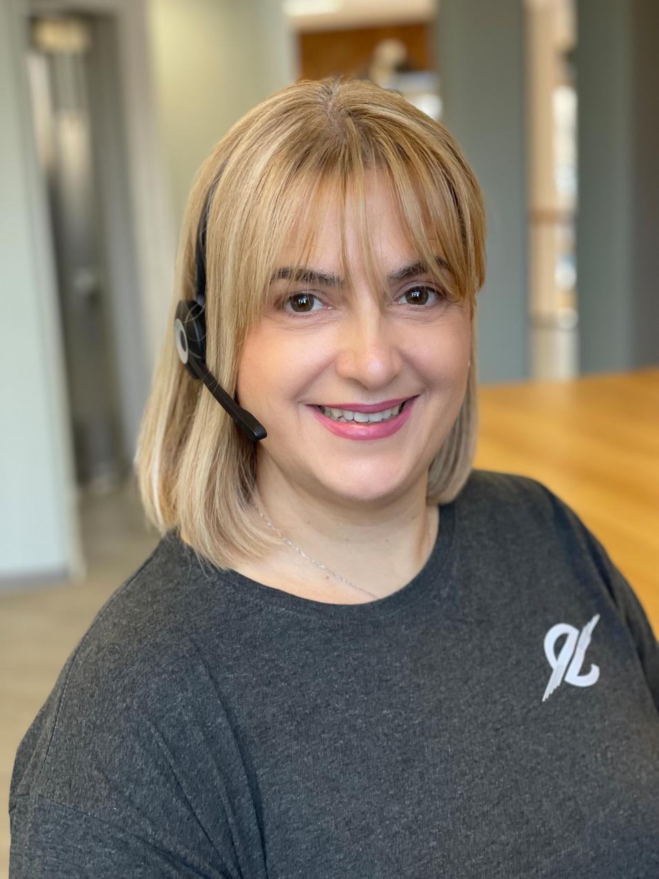 Frau mit kurzer blonder Frisur und Headset l&auml;chelt vor einem modernen Arbeitsplatz.