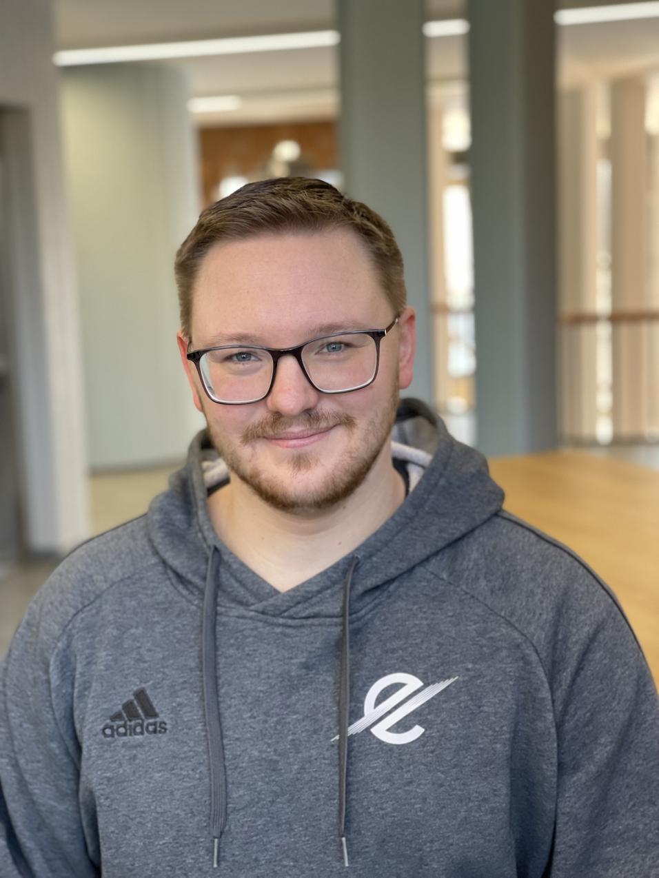L&auml;chelnder Mann mit Brille, grauem Kapuzenpullover und Adidas-Logo, steht in einem modernen Raum mit Holzfu&szlig;boden.
