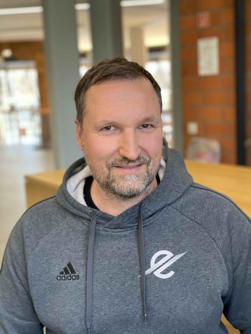 Portr&auml;t eines l&auml;chelnden Mannes mit kurzem Bart, der einen grauen Hoodie tr&auml;gt, vor einer hellen, modernen Umgebung.