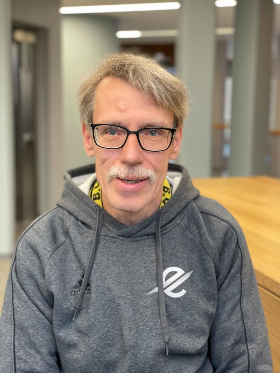 Mann mit Brille und Schnurrbart sitzt in einem hellen Raum, tr&auml;gt grauen Hoodie und schaut freundlich in die Kamera.