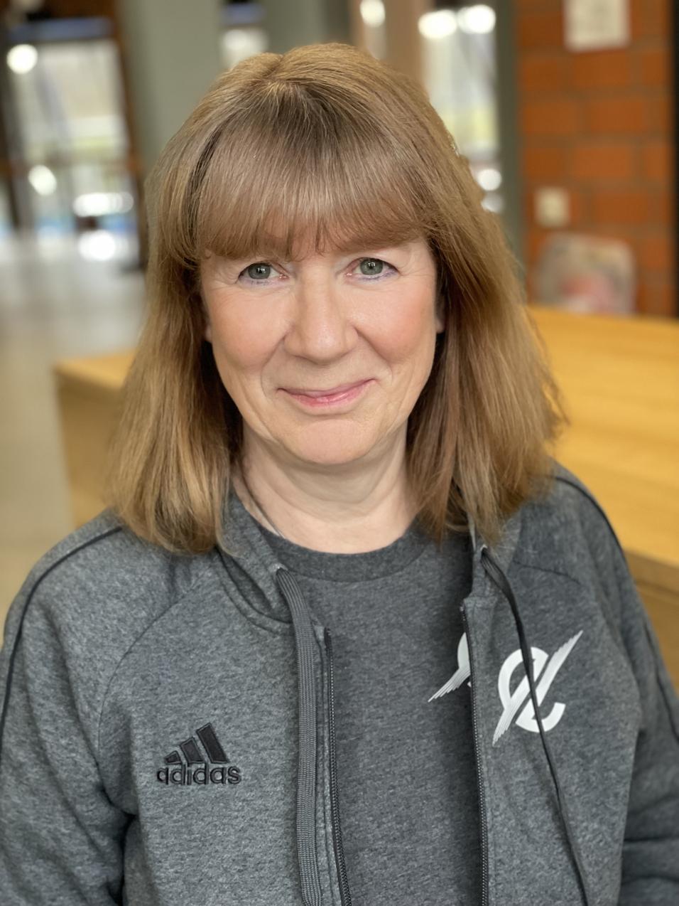 Portr&auml;t einer l&auml;chelnden Frau mit blonden Haaren, die einen grauen Adidas-Trainingsanzug tr&auml;gt, in einem hellen Raum.