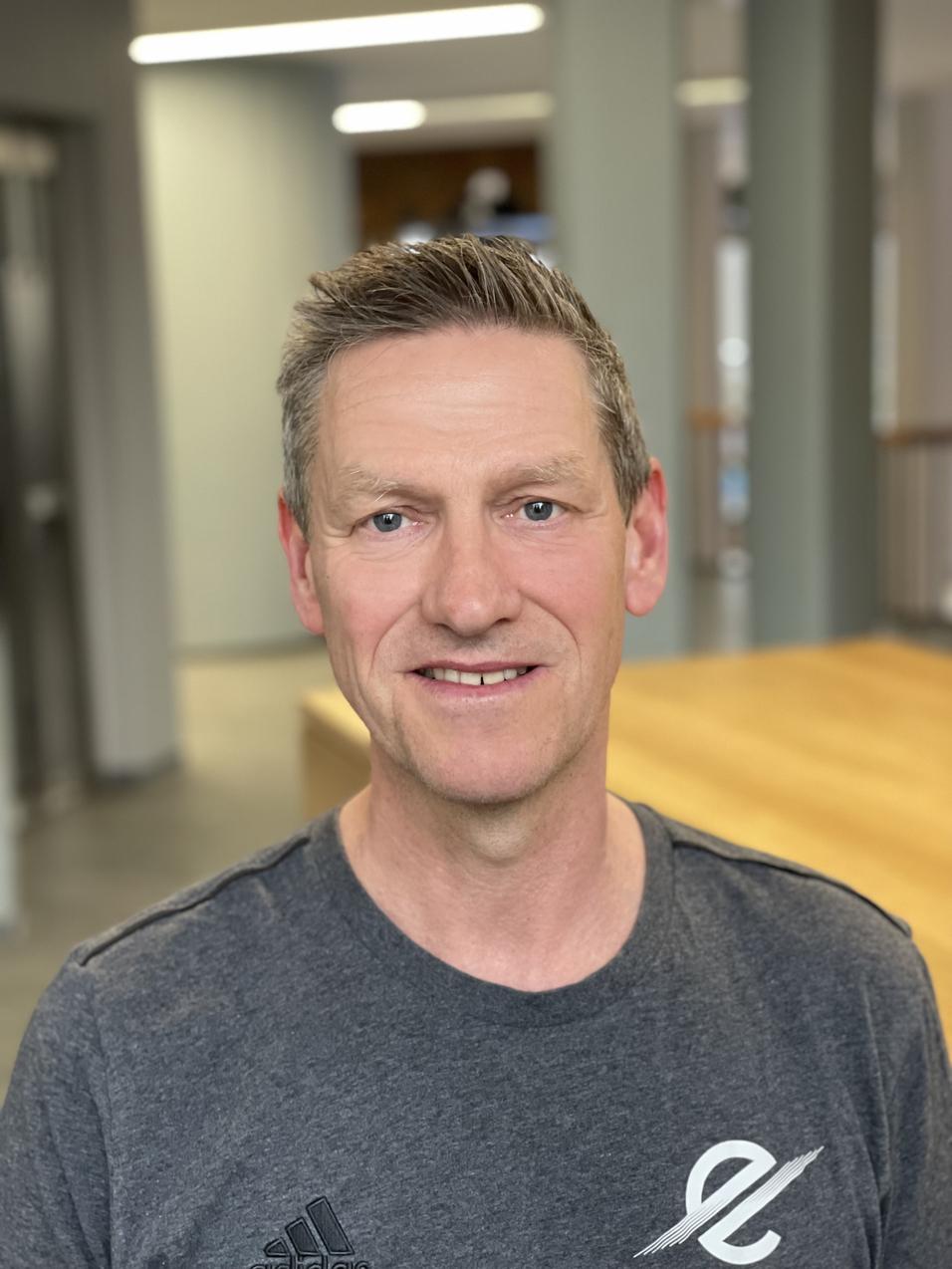 Portr&auml;t eines Mannes mit kurzem, grauen Haar, der in einem sportlichen, grauen T-Shirt in einem modernen Raum l&auml;chelt.