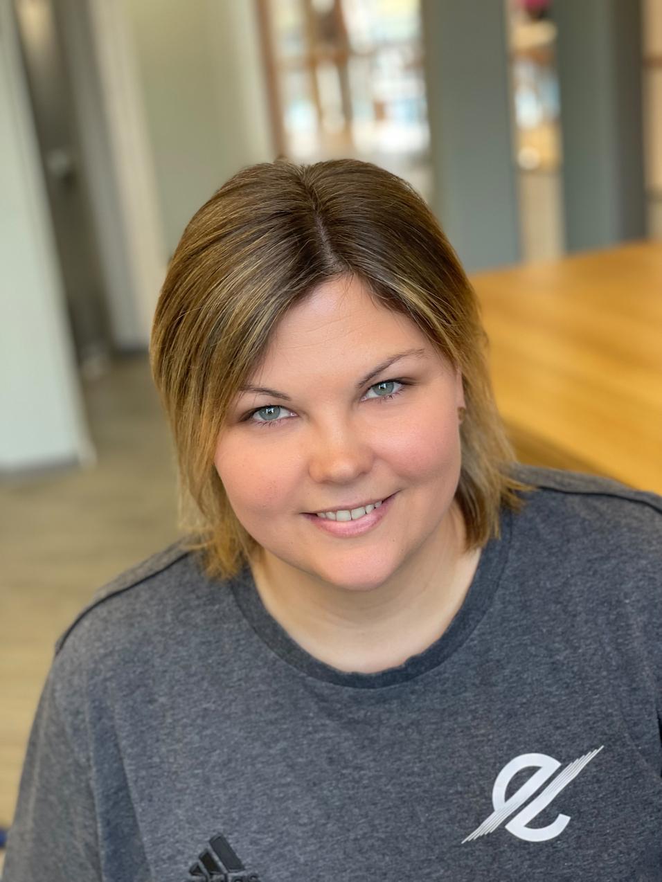 Frau mit blonden Haaren und grauem Shirt l&auml;chelt in einem hellen Raum, mit Holzoberfl&auml;che im Hintergrund.