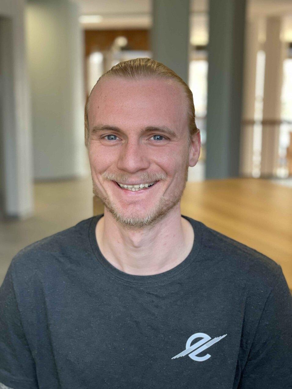 Portrait eines l&auml;chelnden Mannes mit blonden Haaren, der ein schwarzes T-Shirt tr&auml;gt, in einem hellen Innenraum.