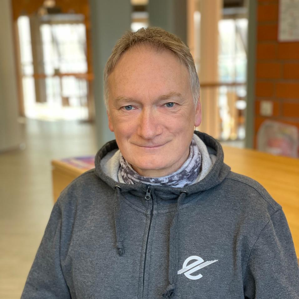 Mann in grauem Hoodie mit Logo steht in einem hellen Innenraum vor einem Holztisch.