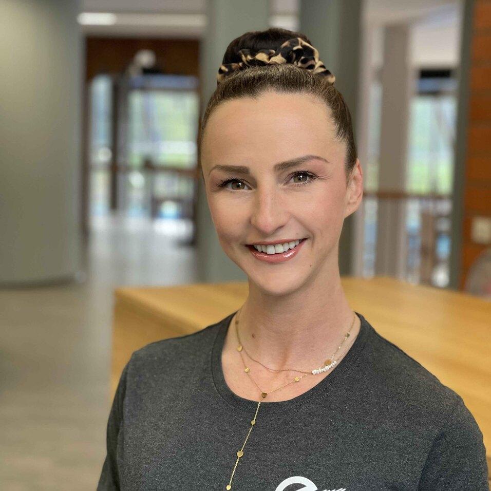 Portr&auml;t einer Frau mit hochgestecktem Haar, die l&auml;chelt. Sie tr&auml;gt ein graues T-Shirt mit einem Logo. Hintergrund verschwommen.