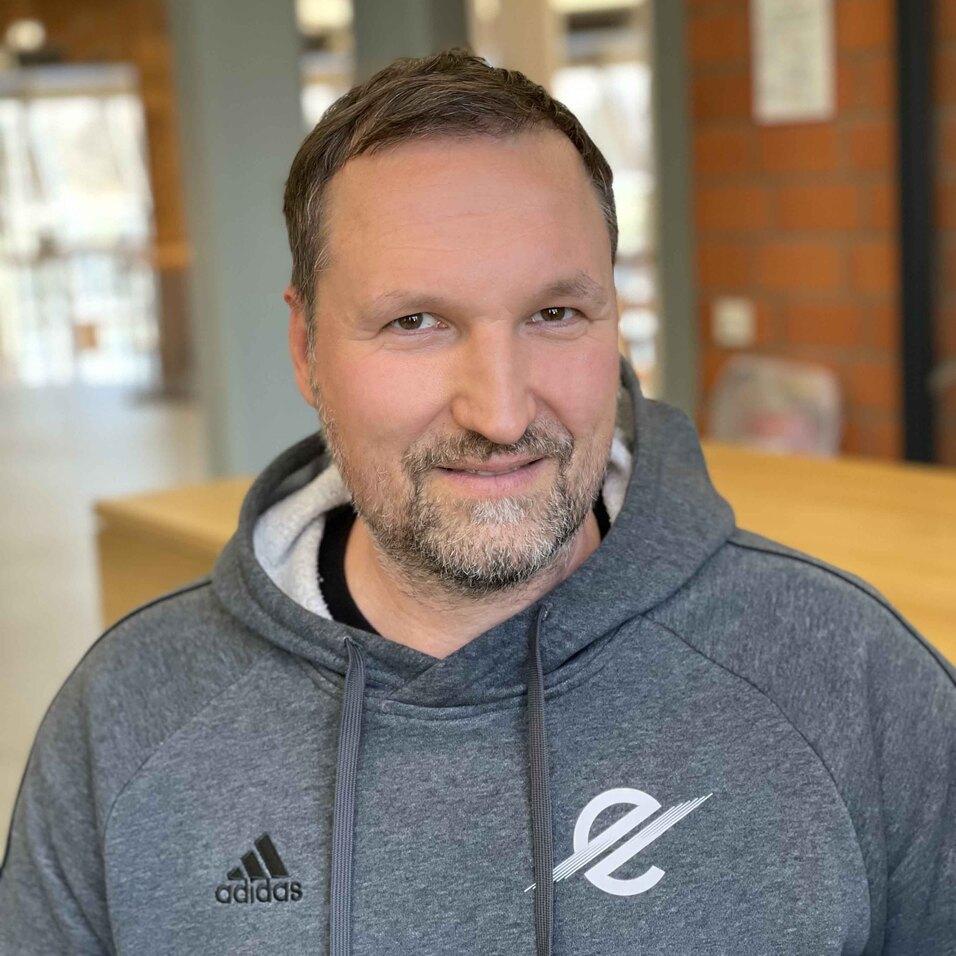 Porträt eines lächelnden Mannes mit kurzem Bart, der einen grauen Hoodie trägt, vor einer hellen, modernen Umgebung.