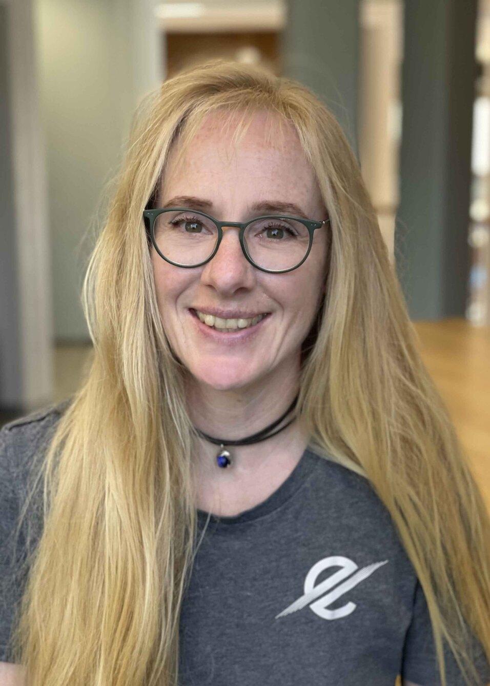 Frau mit langen blonden Haaren und Brille l&auml;chelt vor unscharfem Hintergrund, tr&auml;gt ein graues T-Shirt mit Logo.