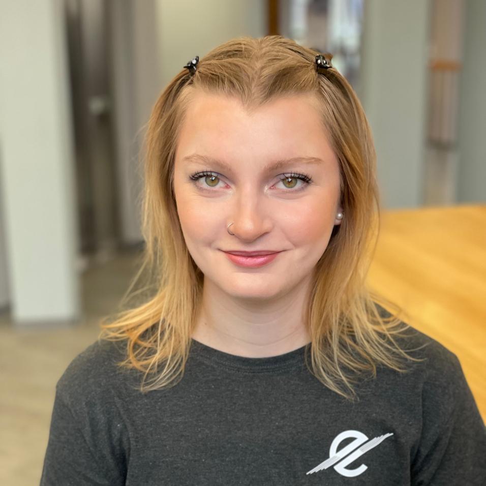 Portrait einer lächelnden Frau mit langen blonden Haaren und einem grauen T-Shirt, sitzend vor einem Holzschreibtisch.