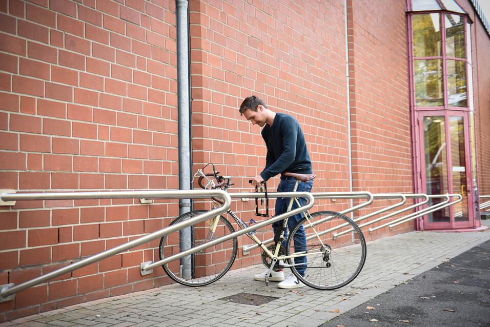 Mann sperrt sein Fahrrad an einer Metallhalterung vor einer Backsteinmauer an.