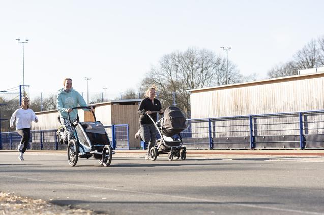 Zwei joggende Erwachsene schieben Kinderwagen auf einer asphaltierten Strecke, w&auml;hrend ein weiterer L&auml;ufer im Hintergrund l&auml;uft.