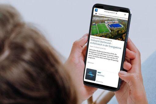 Person hält ein Smartphone, auf dem eine Webseite mit einem Fußballfeld und Text angezeigt wird.
