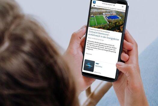 Person hält ein Smartphone, auf dem eine Webseite mit einem Fußballfeld und Text angezeigt wird.