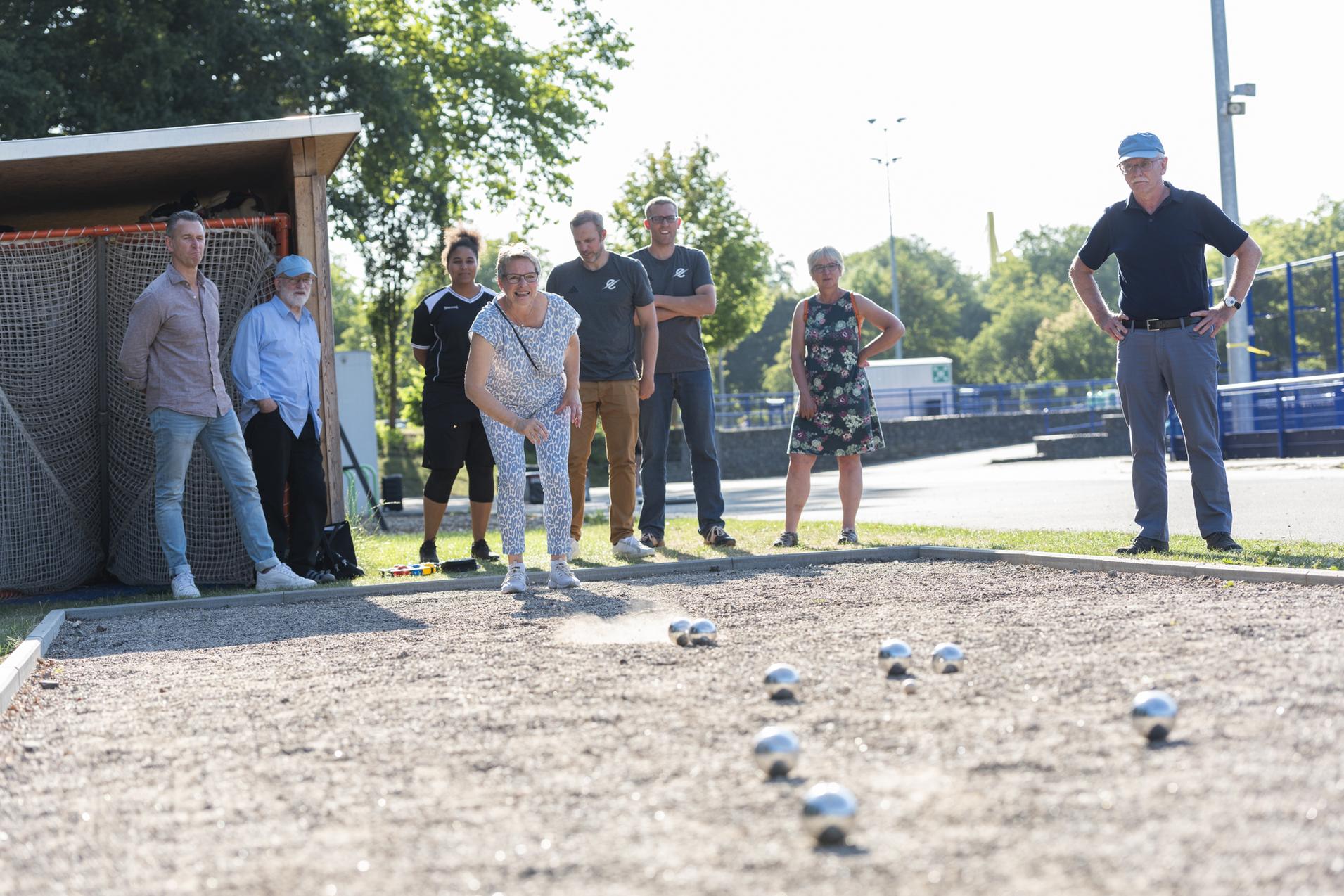 Eine Spielerin wirft eine Metallkugel auf eine Boule-Bahn, w&auml;hrend Zuschauer aufmerksam beobachten.