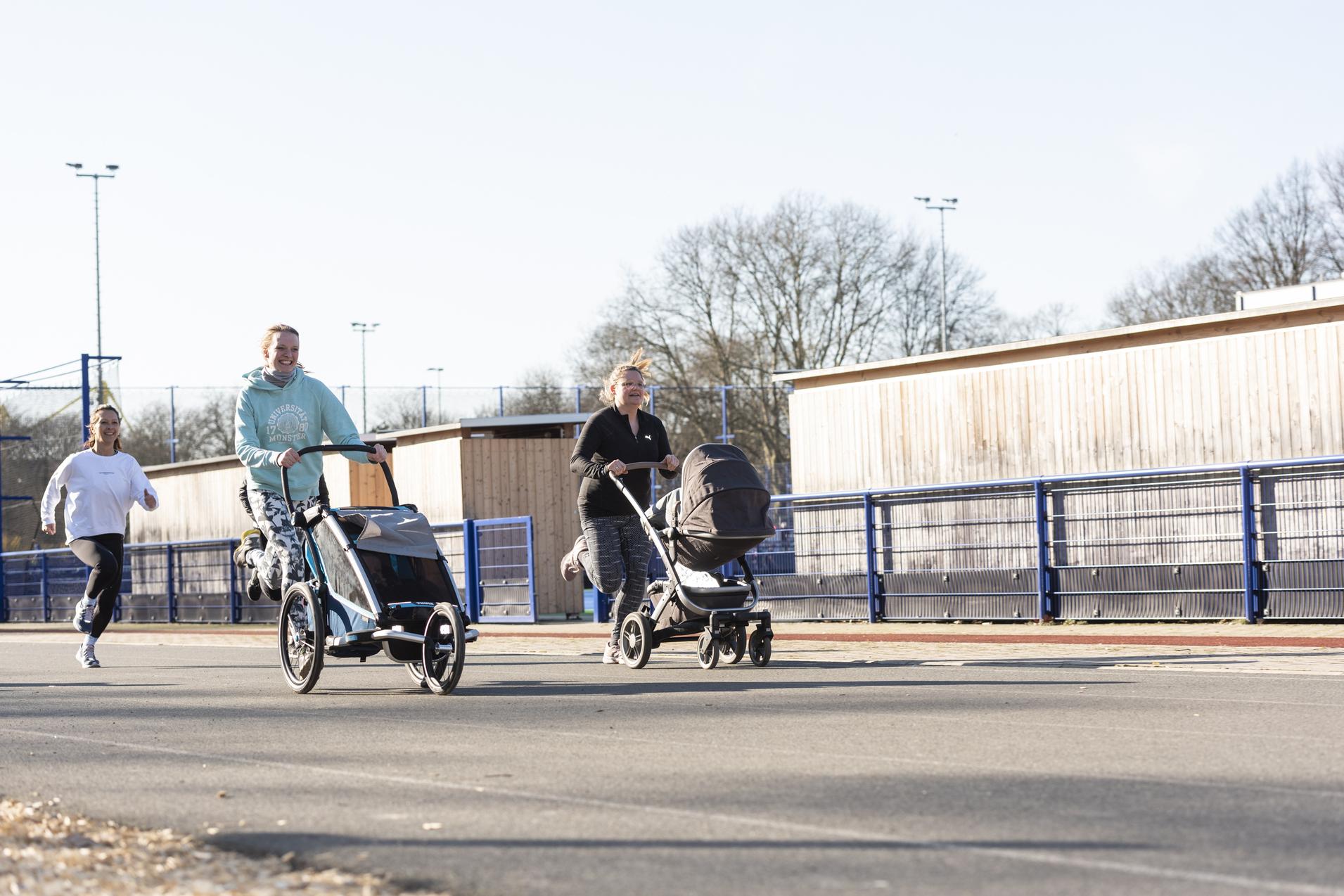 Zwei joggende Erwachsene schieben Kinderwagen auf einer asphaltierten Strecke, w&auml;hrend ein weiterer L&auml;ufer im Hintergrund l&auml;uft.