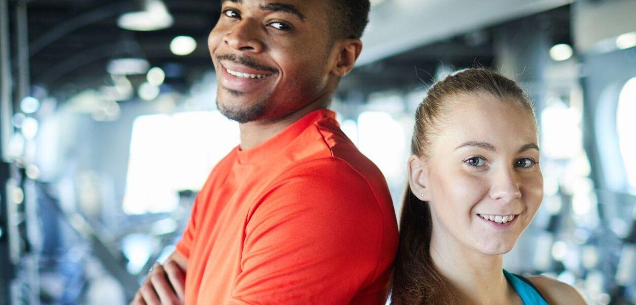Zwei junge Sportler stehen R&uuml;cken an R&uuml;cken in einem Fitnessstudio und l&auml;cheln in die Kamera.