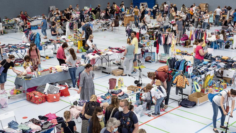 Überblick über eine belebte Sporthalle mit Ständen, an denen Menschen Kleidung und Spielzeug suchen und kaufen.