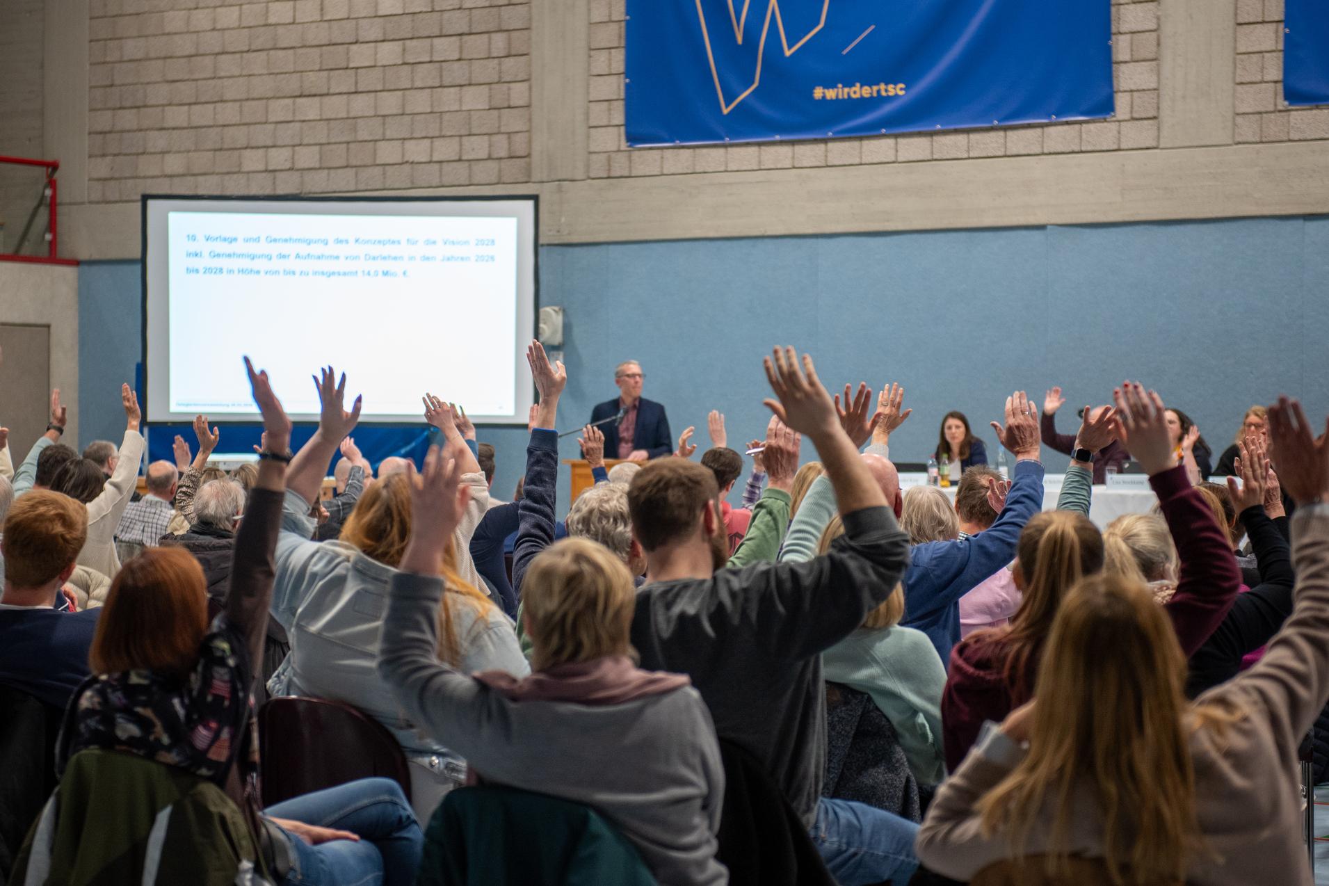Gro&szlig;e Gruppe von Menschen in einem Saal hebt die H&auml;nde w&auml;hrend einer Versammlung. Pr&auml;sentation im Hintergrund sichtbar.