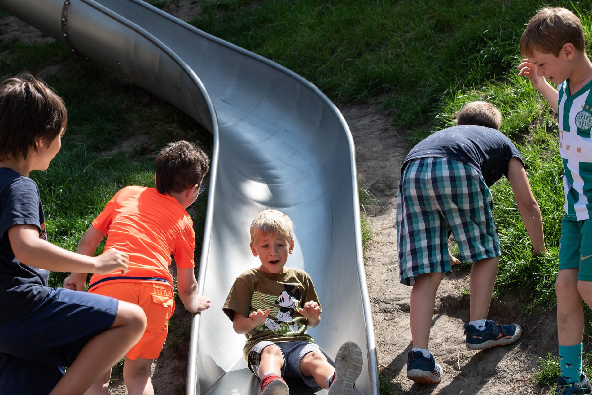 F&uuml;nf Kinder spielen an einer Rutsche, w&auml;hrend einer auf der Rutsche sitzt und die anderen sie umgeben.