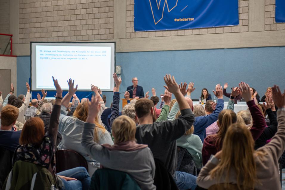 Gro&szlig;e Gruppe von Menschen in einem Saal hebt die H&auml;nde w&auml;hrend einer Versammlung. Pr&auml;sentation im Hintergrund sichtbar.