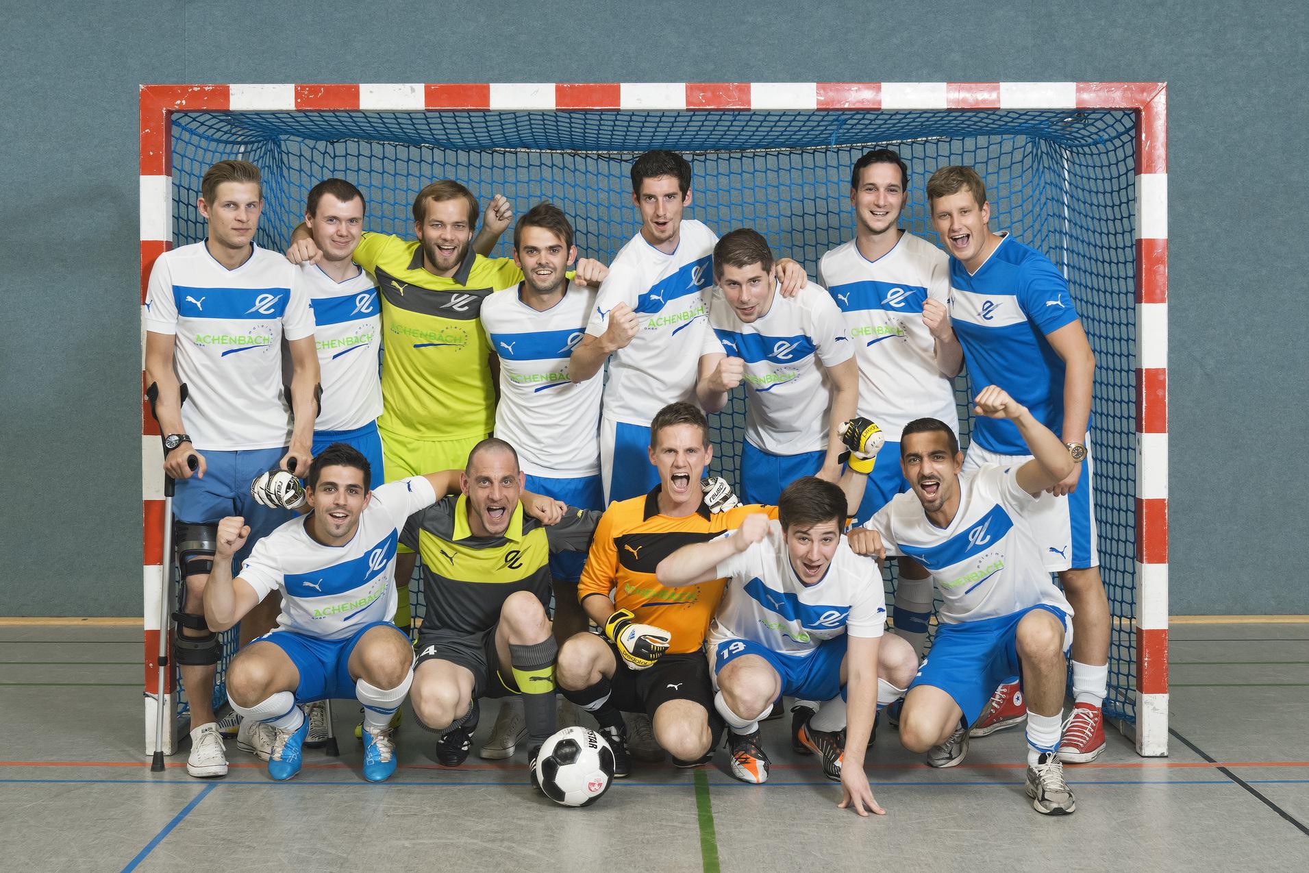 Gruppe von 13 Fußballspielern in blauen und weißen Trikots posiert lächelnd vor einem Tor in einer Sporthalle.